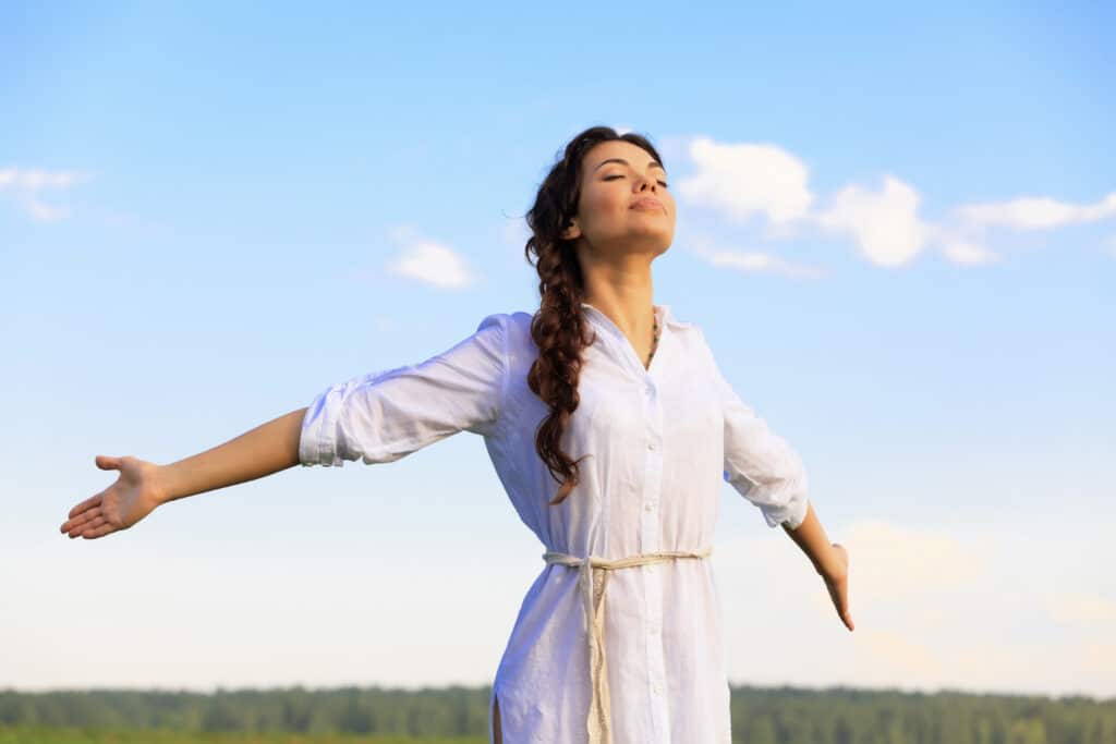 Ung kvinde med åbne arme og lukkede øjne indånder frisk luft i naturen med en blå himmel i baggrunden